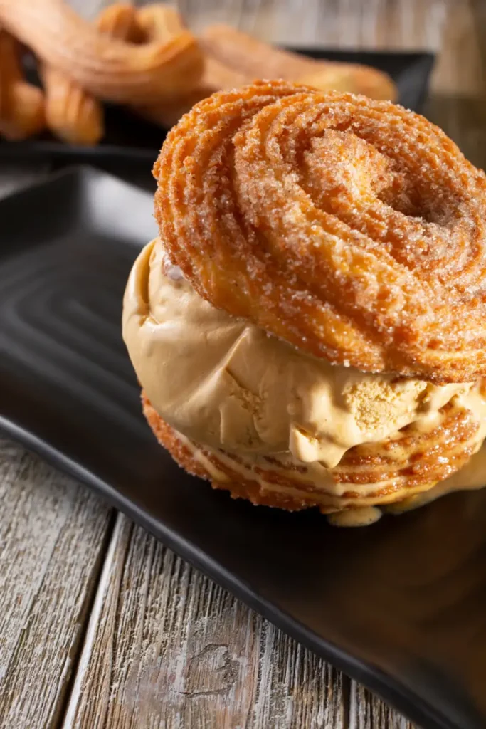 A churro ice cream sandwich with caramel-flavored ice cream on a black rectangular plate, with another churro in the background, all on a wooden surface.