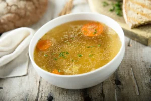 A bowl of chicken bouillon garnished with chopped parsley, containing slices of carrot, served on a rustic wooden table with whole grain bread and a linen napkin.