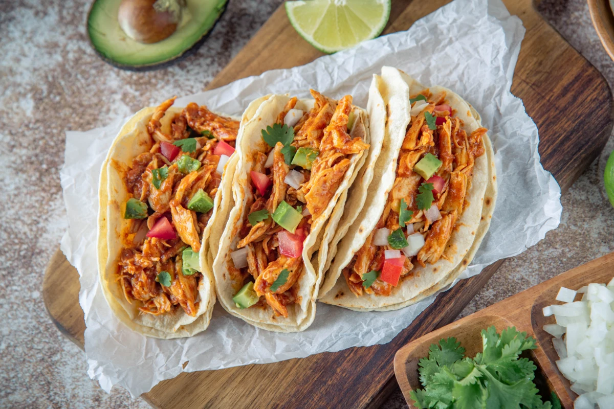 Three chicken tacos filled with shredded chicken, diced tomatoes, chopped avocados, and cilantro on soft tortillas, served on crumpled white paper. Accompaniments include sliced lime and half an avocado in the background, with diced onions and a bunch of cilantro on the side.