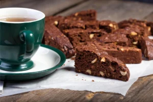 A green cup of tea with saucer on a wooden surface next to chocolate biscotti pieces with visible nuts.