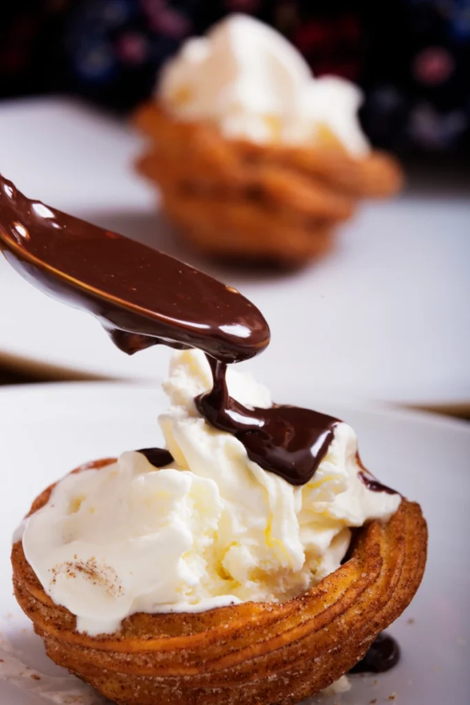 Chocolate sauce being poured over a churro ice cream sandwich with vanilla ice cream, on a white plate against a blurred background of berries.