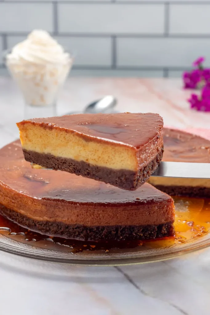 A close-up of a chocolate flan cake with a glossy caramel topping and a single slice lifted, ready to be served. In the background, a whipped cream topping in a glass and purple flowers add a decorative touch to the scene.