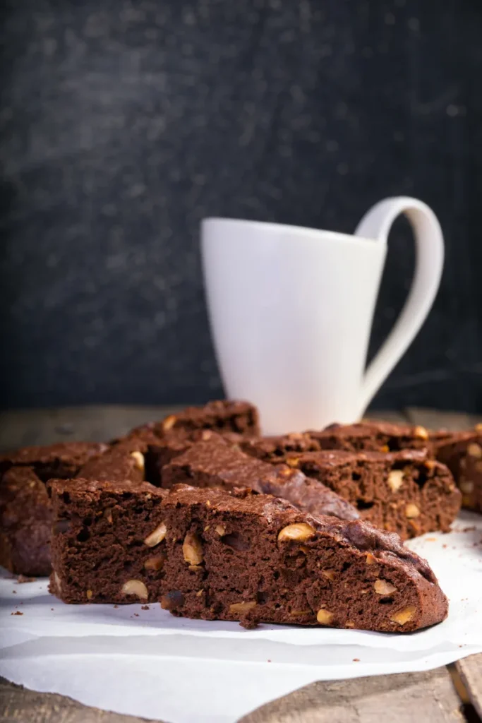 Sliced chocolate biscotti with nuts on parchment paper, with a white mug in the soft-focused background, against a dark textured backdrop.