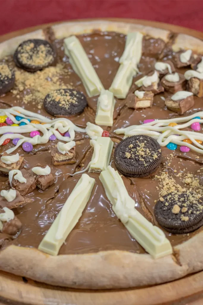 A dessert pizza with chocolate spread base, topped with white chocolate bars arranged in a star pattern, cookie pieces, and colorful candy-coated chocolates.