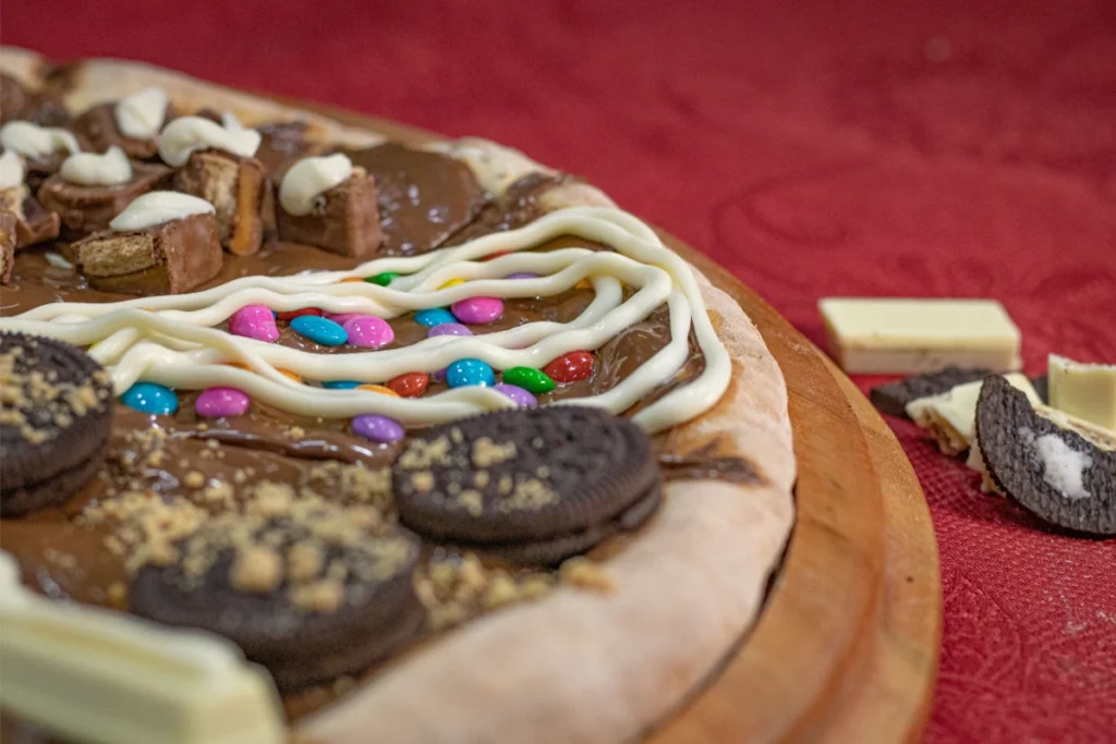 A close-up view of a homemade chocolate pizza on a wooden board with a variety of sweets as toppings, including colorful candy-coated chocolates, chocolate-filled cookies, and chocolate pieces, against a red fabric background.