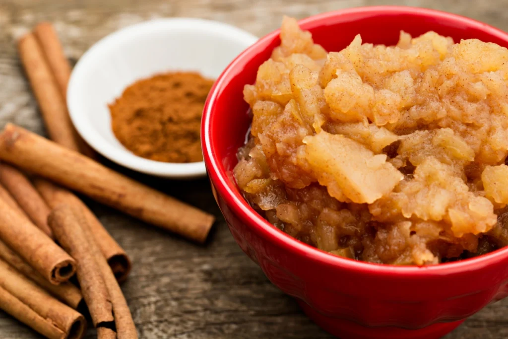 A red bowl of chunky cinnamon applesauce on a wooden surface, with cinnamon sticks and a small bowl of ground cinnamon in the background.