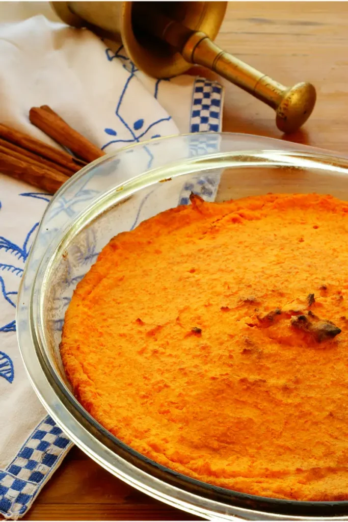 Golden-brown carrot soufflé in a clear glass pie dish, placed on a blue and white checked cloth next to cinnamon sticks and a brass mortar and pestle on a wooden table.