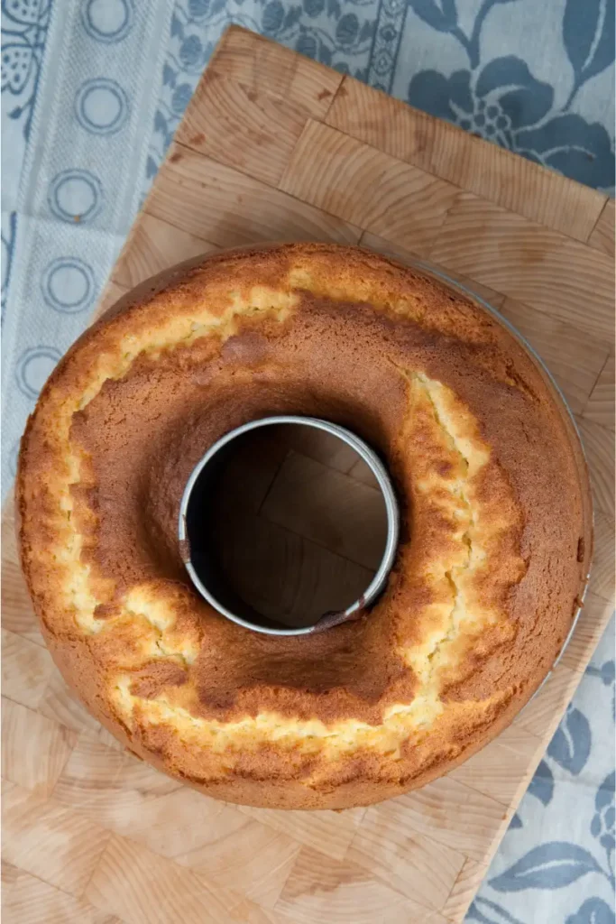 Overhead view of an uncut Bishop Cake on a wooden cutting board, displaying a golden-brown crust, with a blue patterned textile partially visible in the background.