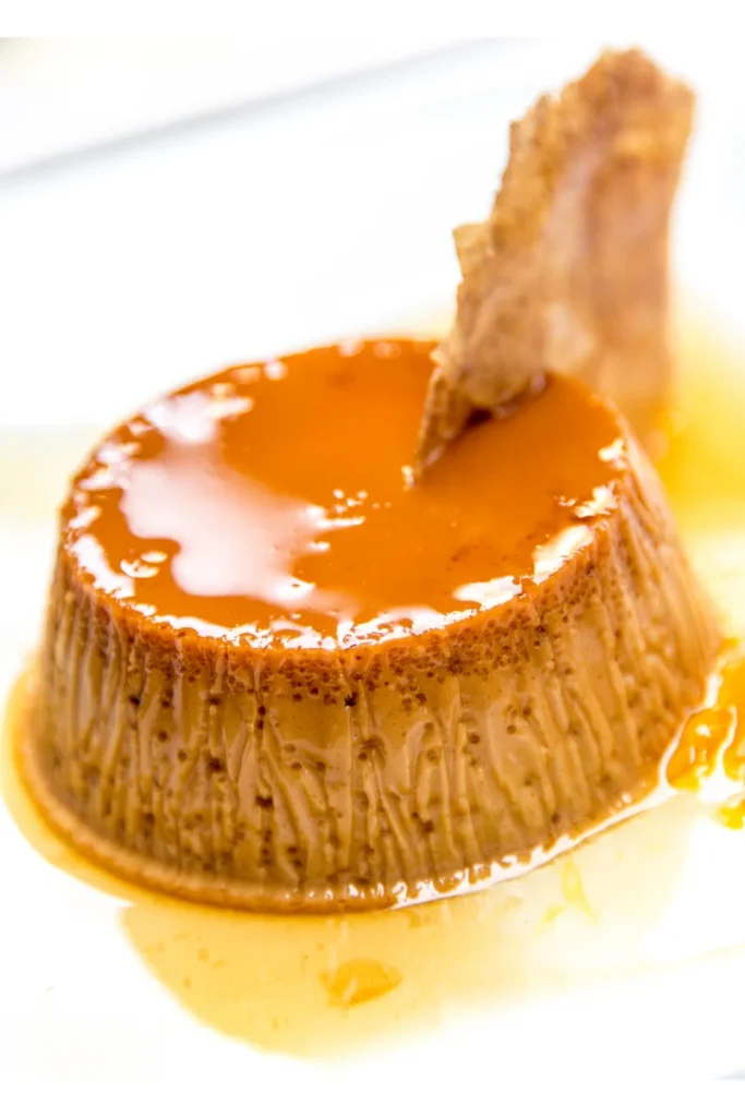 A coffee flan with a shiny caramel glaze on top and liquid caramel pooling around it, with a piece of hardened caramel inserted at an angle into the flan.