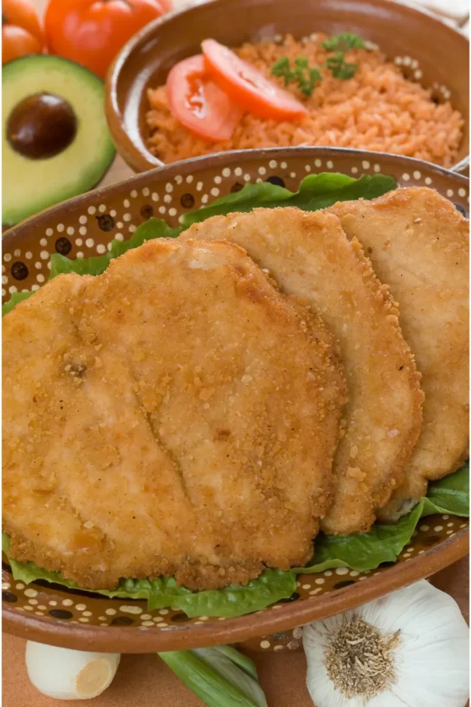 Breaded milanesa steaks served on a bed of lettuce in a decorative clay plate, accompanied by avocado, tomato, and seasoned rice.