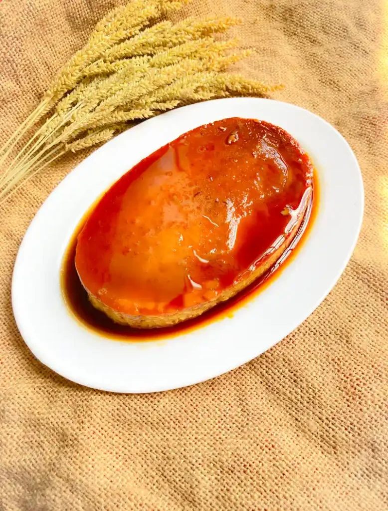 A single pumpkin flan with caramel topping on a white plate, placed on a textured burlap cloth with wheat stalks to the side.