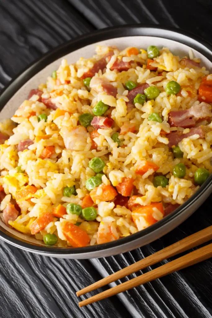 A bowl of Yangzhou Fried Rice with green peas, carrot cubes, scrambled eggs, and ham, accompanied by wooden chopsticks, on a dark wooden table.