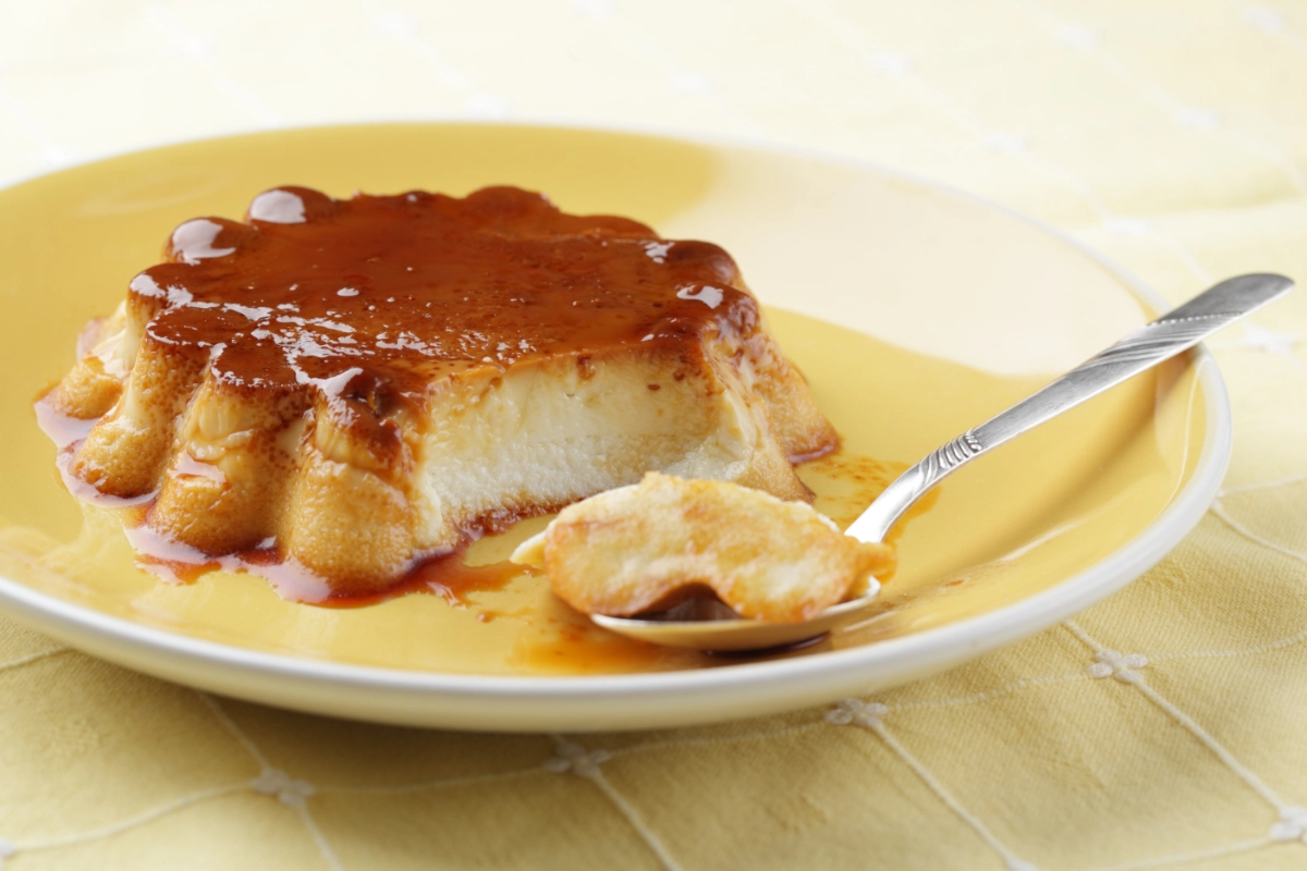 A slice of coconut flan with caramel topping on a yellow plate, with a fork taking a piece, on a textured yellow background.