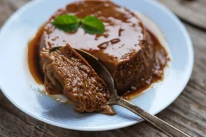 A close-up of a coffee flan on a white plate with blue stripes, garnished with a fresh basil leaf, with caramel sauce drizzled over and around the dessert. A spoon is taking a portion out, revealing a smooth, creamy texture.