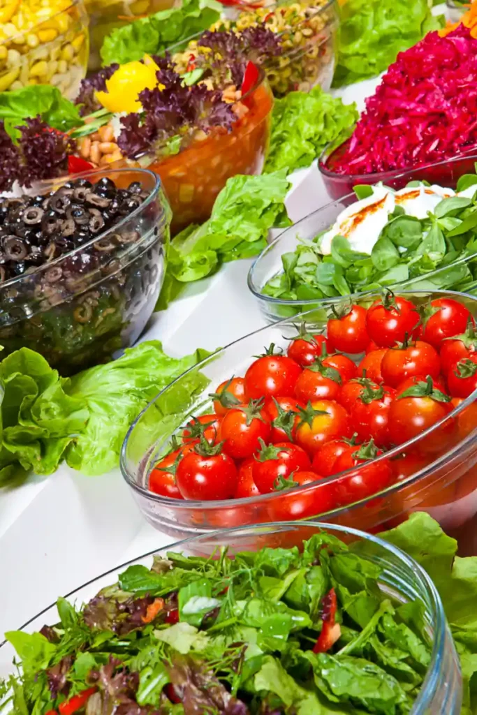 Assorted fresh salads displayed in clear glass bowls, featuring cherry tomatoes on the vine, sliced black olives, golden corn kernels, shredded beetroot, various leafy greens, and a mixed salad with nuts.