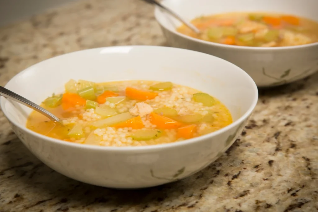 Two bowls of pastina soup with chunks of carrot, celery, and chicken on a granite countertop, one bowl in the foreground with a spoon.