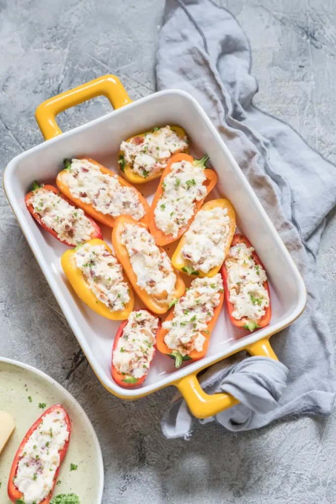 Stuffed bell peppers with cream cheese and herbs in a ceramic baking dish with yellow handles, alongside a gray linen napkin on a gray textured background.