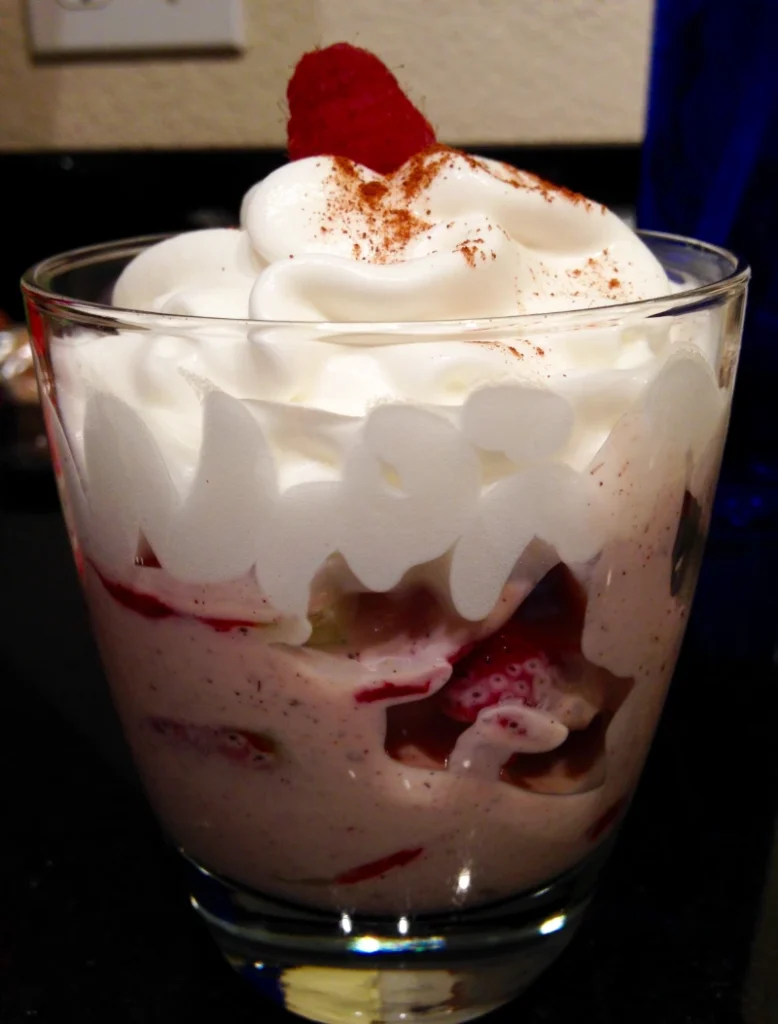 Whipped cream topped strawberry dessert with a dusting of cocoa, garnished with a strawberry in a clear glass.