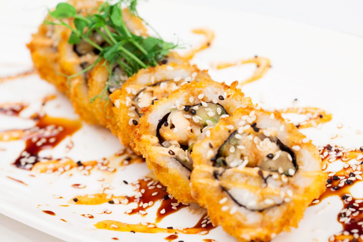 A close-up of a crunchy roll sushi, garnished with green herbs and sprinkled with sesame seeds, presented on a white plate with artistic drizzles of soy sauce.