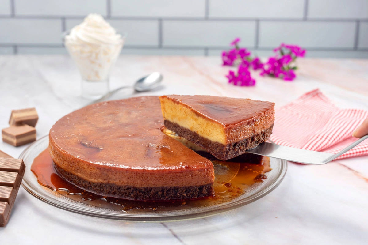 A whole chocolate flan cake with one slice partially removed, revealing its layered structure, on a clear glass plate with caramel sauce. In the background are pieces of chocolate, a white spoon, a glass with whipped cream, a pink striped napkin, and small purple flowers.