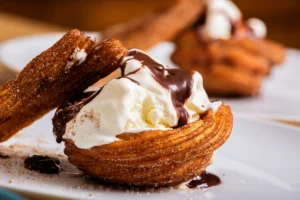 Close-up of a churro ice cream sandwich, with a spiraled churro filled with vanilla ice cream and drizzled with melted chocolate, on a white plate with sugar sprinkled around.