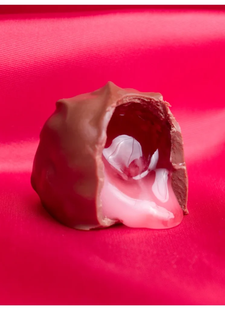 A chocolate-covered cherry with a bite taken out, revealing white creamy filling on a red background.