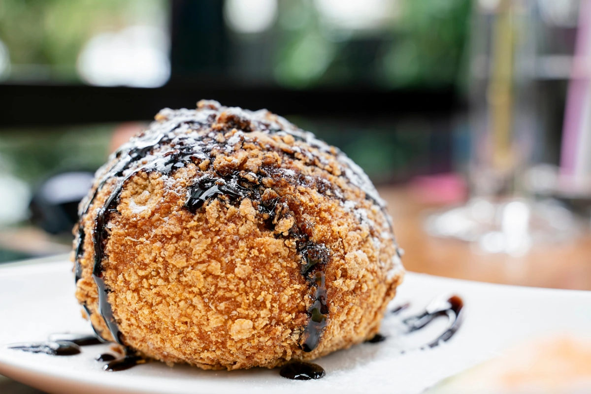 Close-up of a deep-fried ice cream ball drizzled with chocolate syrup, sprinkled with powdered sugar, presenting a crispy golden-brown crust, on a white plate with a blurred background.