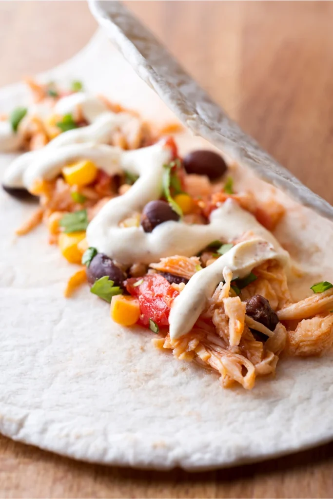 Close-up view of an open chicken taco on a soft tortilla, featuring shredded chicken, black beans, diced tomatoes, corn, green onions, and a drizzle of creamy white sauce.