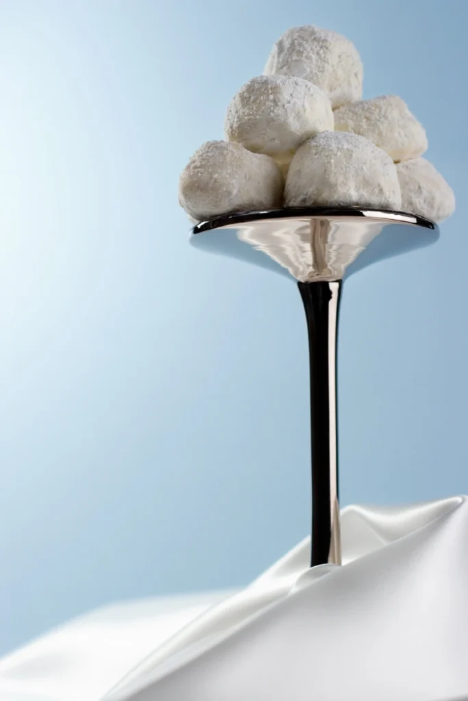 A tall, elegant metal stand on a draped white cloth holding a pyramid of Mexican Wedding Cakes, each dusted with powdered sugar, set against a serene blue backdrop.