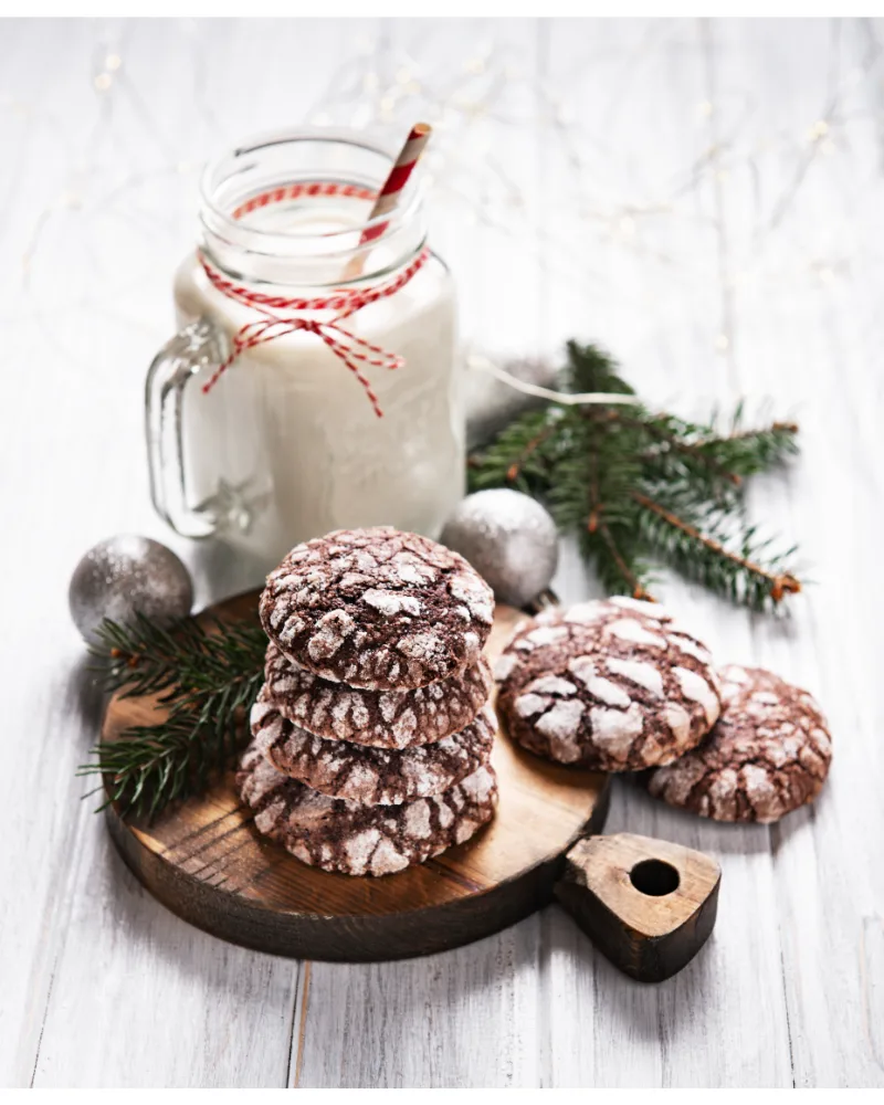 A stack of Cool Whip cookies on a wooden cutting board, with a jar of milk adorned with a festive straw and Christmas decorations in the background.