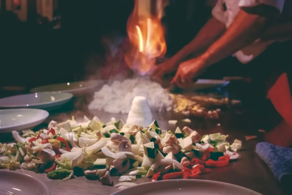 A teppanyaki chef's hands creating a dramatic flame on a grill surrounded by assorted chopped vegetables and rice, with plates awaiting the cooked meal.