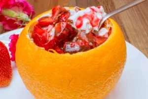 Dessert of strawberries and cream in an orange bowl on a white plate, with a pink flower and strawberries in the background.