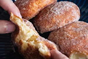 A close-up of sugar-coated malasadas, with one being pulled apart to show the fluffy interior. The pastries exhibit a golden-brown exterior and are presented in a group, with the focus on the texture and sugary coating.