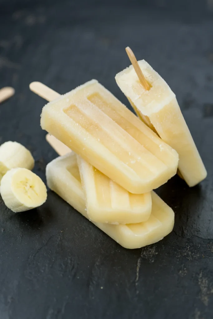 Stack of three banana popsicles with one partially bitten, arranged on a dark slate background, accompanied by sliced bananas to the side.