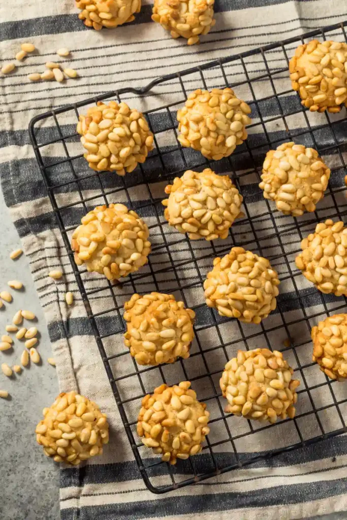 Pignoli cookies with golden brown edges and white pine nuts on top, cooling on a black wire rack over a striped cloth.