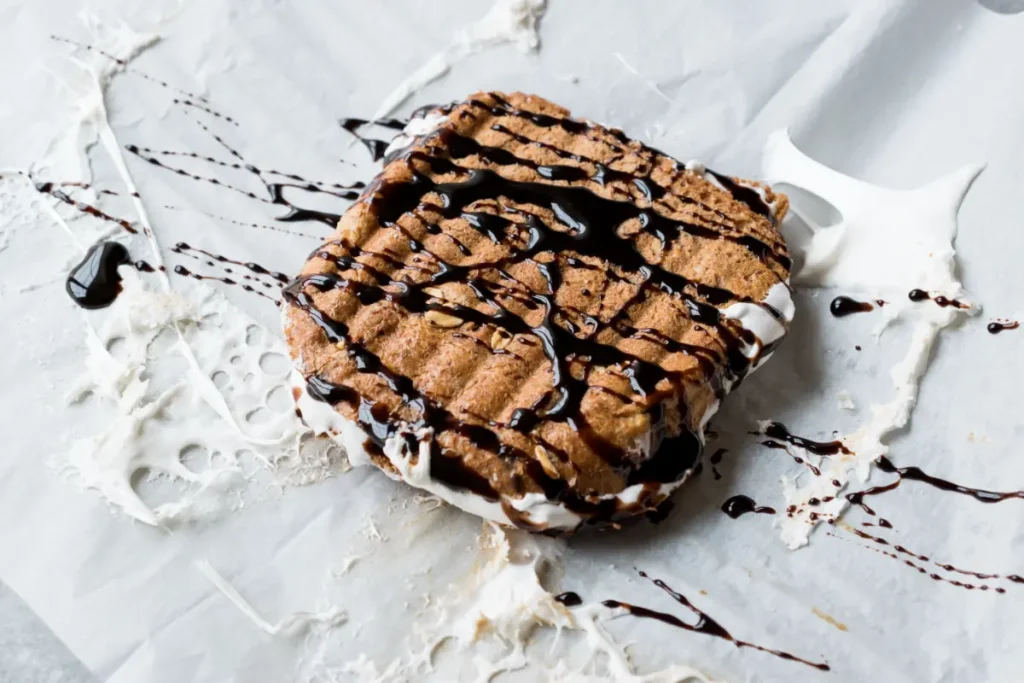 A fluffernutter sandwich with a visible marshmallow filling, topped with a zigzag pattern of chocolate syrup, set against a parchment paper backdrop with syrup drizzles.