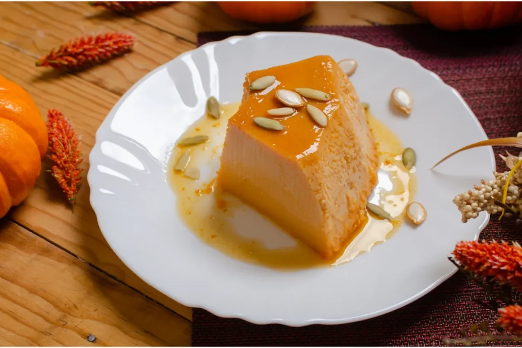 A slice of pumpkin flan topped with pumpkin seeds, on a white plate with caramel sauce, beside fall decorations including an orange pumpkin and dried red flowers.