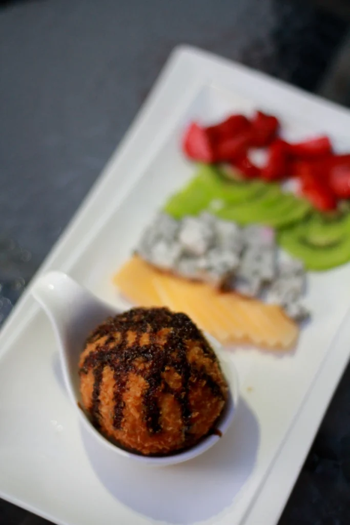 A single scoop of deep-fried ice cream with grill marks, presented in a small white bowl on a rectangular plate, accompanied by sliced strawberries, avocado, and cookies and cream dessert, all on a dark surface.