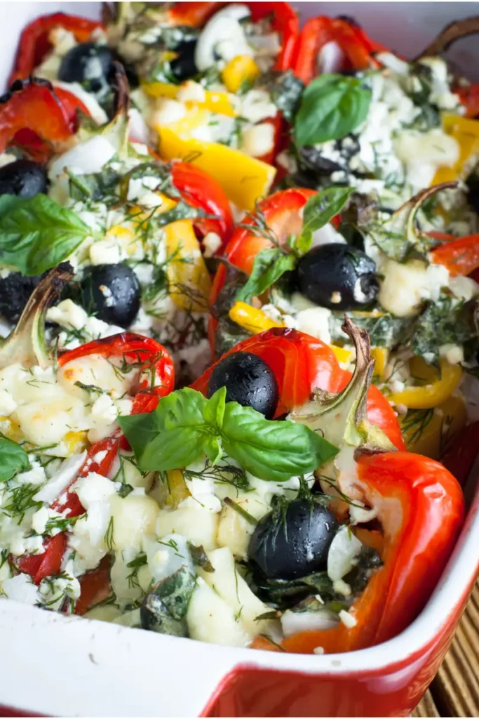 A dish of colorful stuffed bell peppers with cream cheese, black olives, chopped herbs, and melted cheese, garnished with fresh basil leaves.