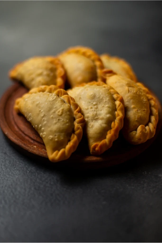 A plate of pumpkin empanadas with a golden, bubbly crust, presented on a wooden plate against a dark background.