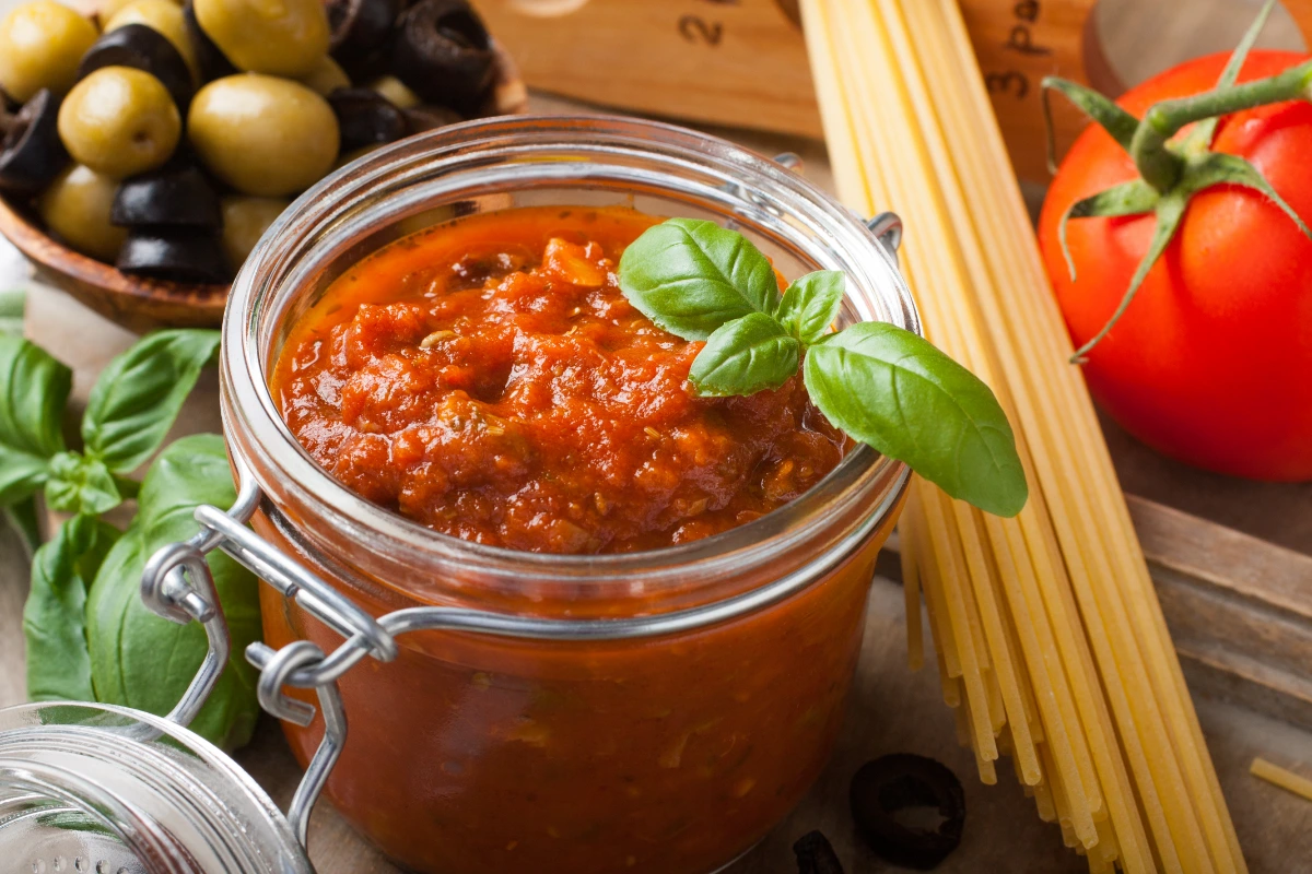 A jar of homemade tomato sauce with fresh basil on top, alongside ingredients like spaghetti pasta, fresh tomato, olives, and basil leaves.