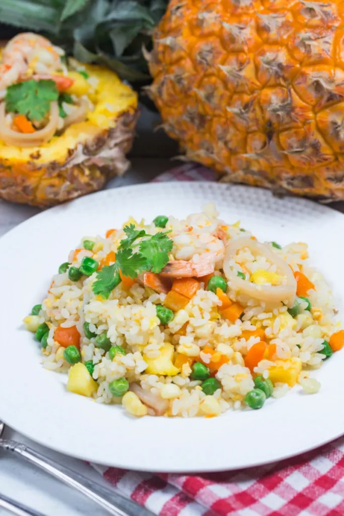 A plate of pineapple fried rice with shrimp, peas, and carrot pieces, garnished with fresh cilantro. In the background, a whole pineapple and a red-checkered napkin complete the tropical presentation.