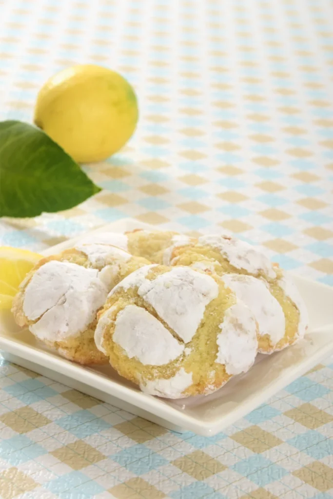 Powdered sugar-dusted Cool Whip cookies on a white plate, with a fresh lemon and leaf in the background, placed on a checkered blue and white tablecloth.