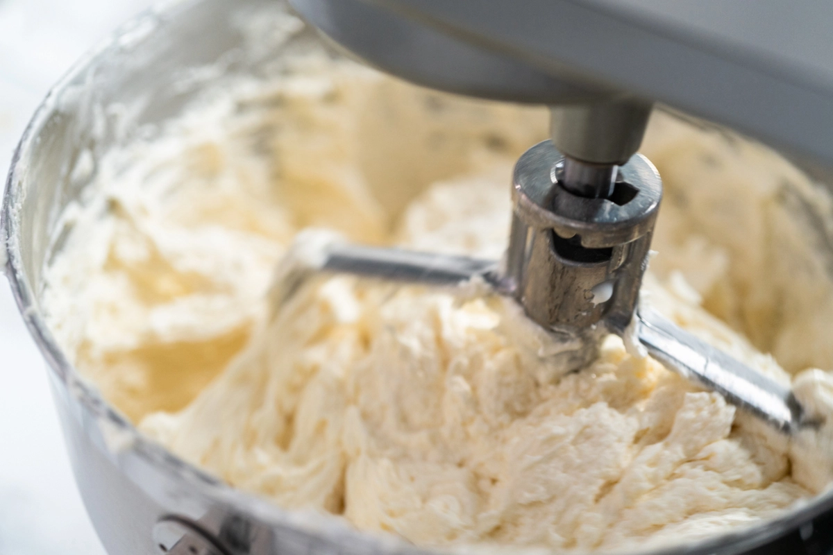 Close-up of a metal whisk attachment mixing Russian buttercream in a stainless steel bowl.