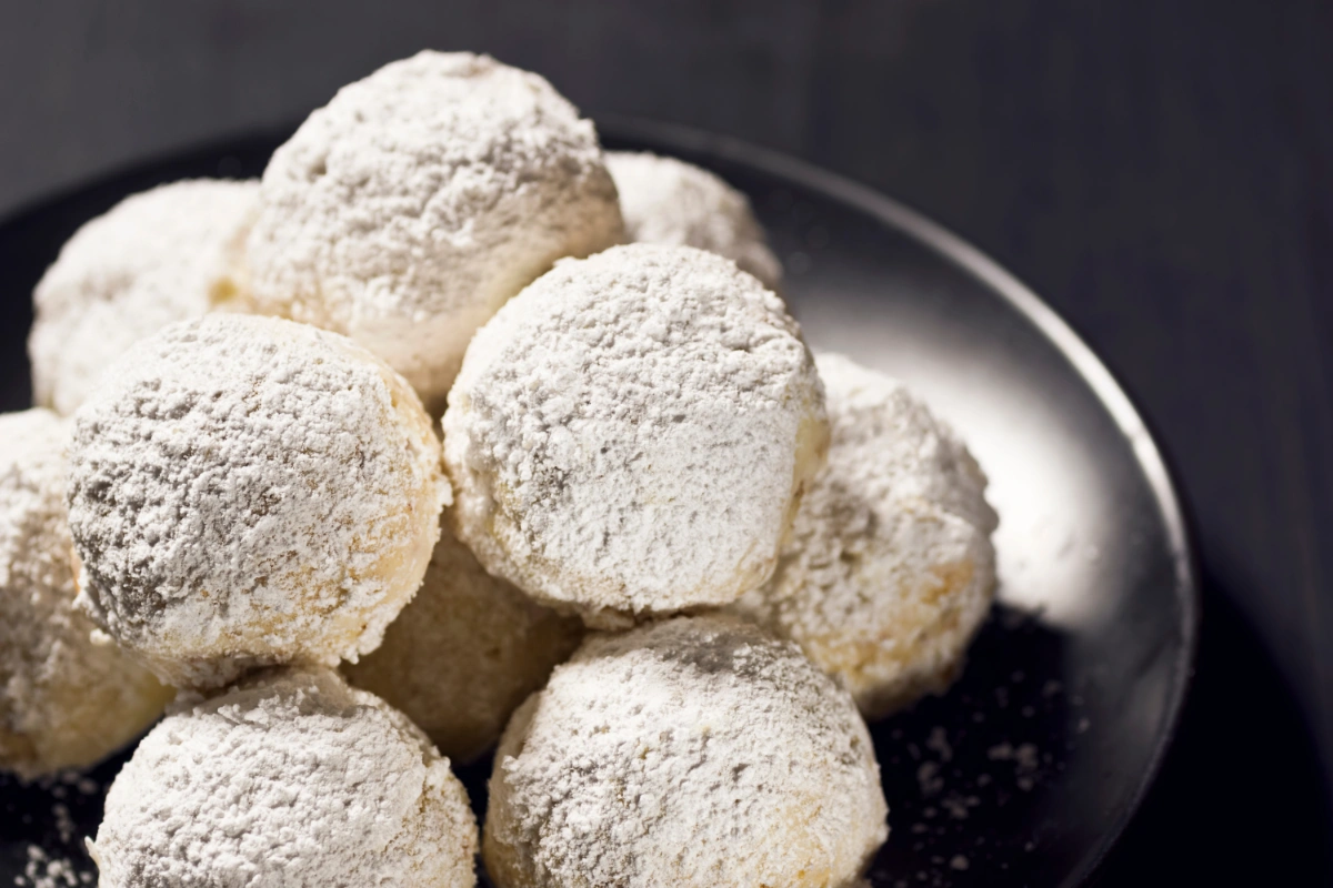 A close-up view of Mexican Wedding Cakes, round powdered sugar-coated cookies, on a dark plate against a dark background.