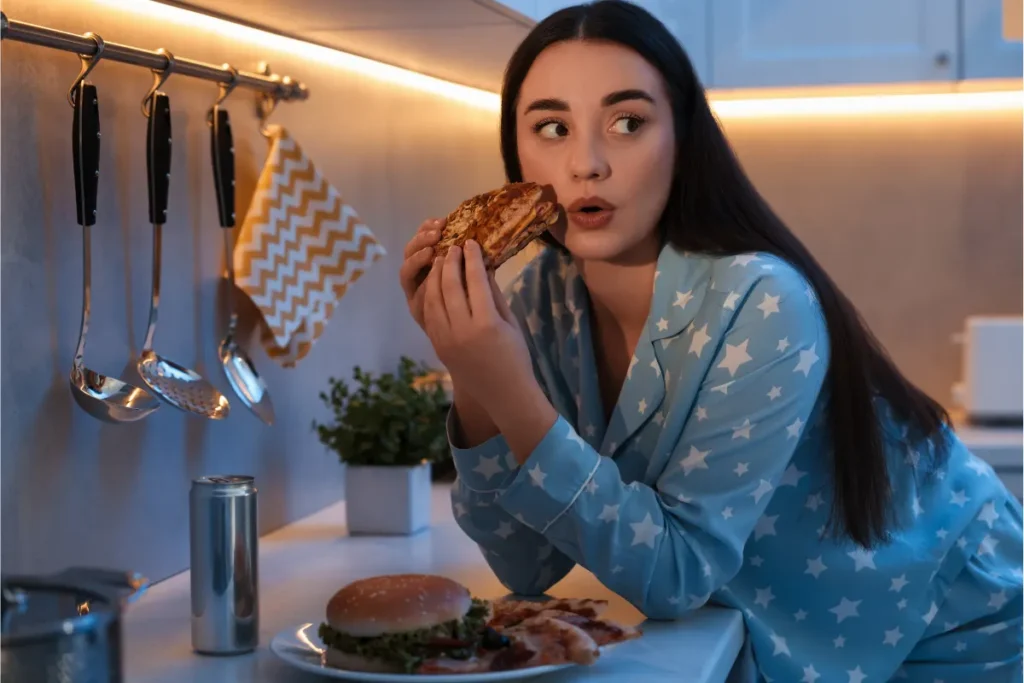 A surprised woman in star-patterned pajamas eating a grilled sandwich in a dimly lit kitchen with a burger and a can on the countertop.