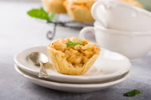 A mini apple pie with a lattice crust on a small plate next to a silver spoon, with more pies and a white tureen in the background, on a grey countertop.