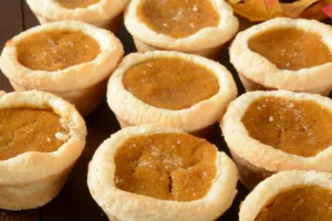 A group of mini pumpkin pies with golden-brown filling, sprinkled with granulated sugar, displayed on a wooden surface.