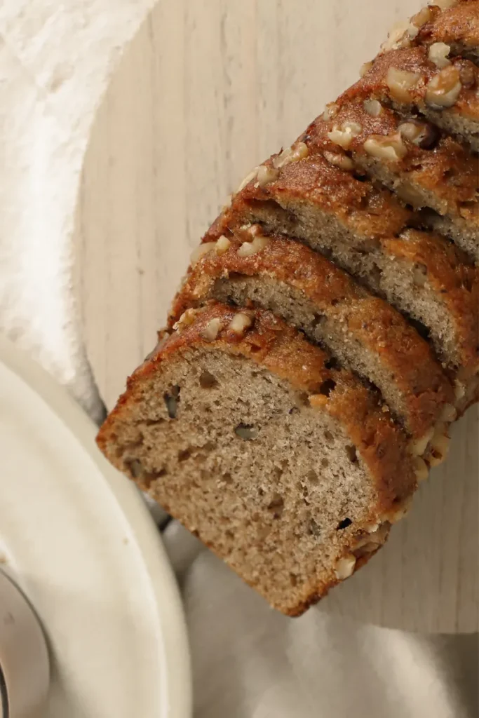 Close-up of vegan banana bread with a crunchy walnut topping, sliced and resting on a light wood surface, showcasing its soft texture and speckled banana pieces.