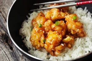 Bowl of Panda Express Honey Sesame Chicken served on a bed of white rice, garnished with sesame seeds and green onion, with chopsticks on the side.